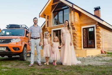Family of four happily standing outside their modern wooden house on a sunny day.