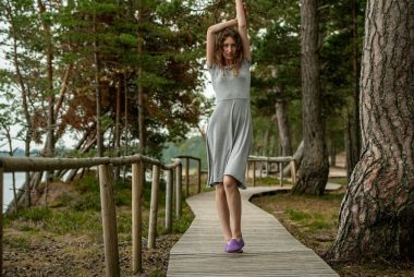 A slim dark-haired woman in a gray dress walking along a wooden boardwalk by the sea, surrounded by coastal forest.